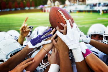 football players huddles in a circle reaching for the football above